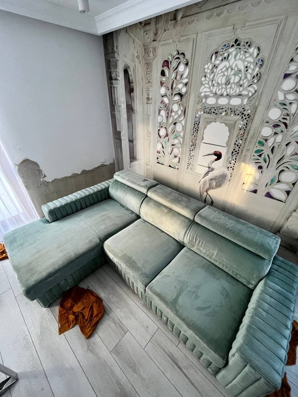 Turkusowy, welurowy narożnik z dekoracyjnym pikowaniem na tle ściany z motywem architektonicznym i wizerunkiem żurawia, leżąca na podłodze brązowa szmatka.