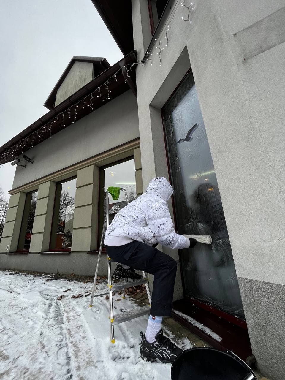 Osoba w białej kurtce na drabinie czyści okno budynku z dekoracjami świątecznymi, widoczny śnieg na ziemi.