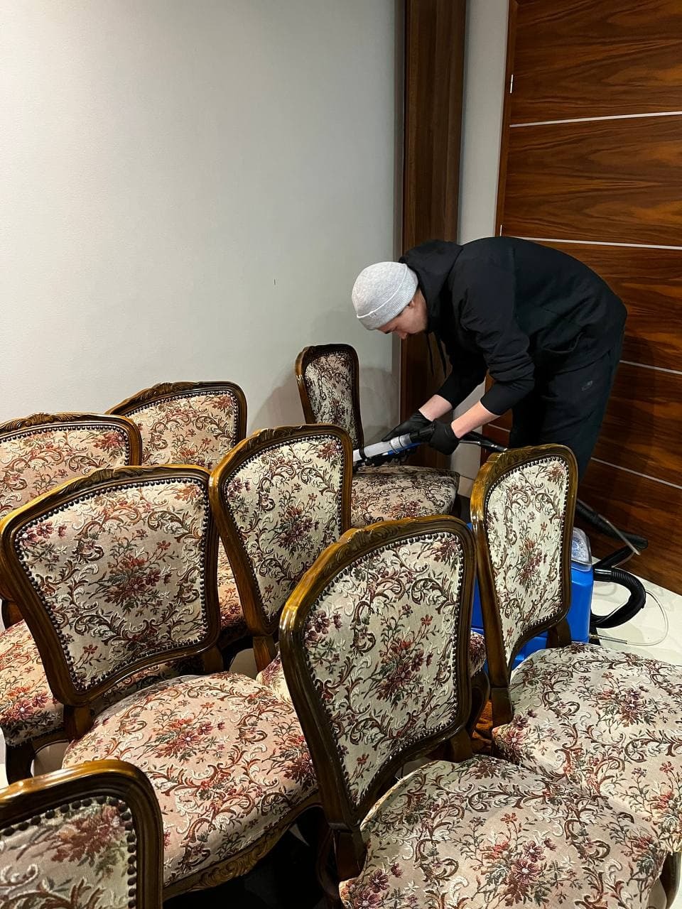 Pracownik w czarnej bluzie i czapce, w czarnych rękawicach, czyści tapicerkę krzesła z florystycznym wzorem za pomocą odkurzacza piorącego. W tle widoczne inne krzesła z podobną tapicerką...