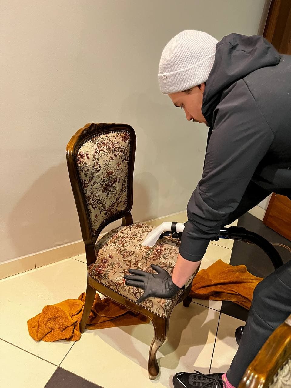 Osoba w czarnych rękawiczkach czyści tapicerkę krzesła z wzorem kwiatowym za pomocą odkurzacza piorącego. Na podłodze leży pomarańczowy ręcznik.