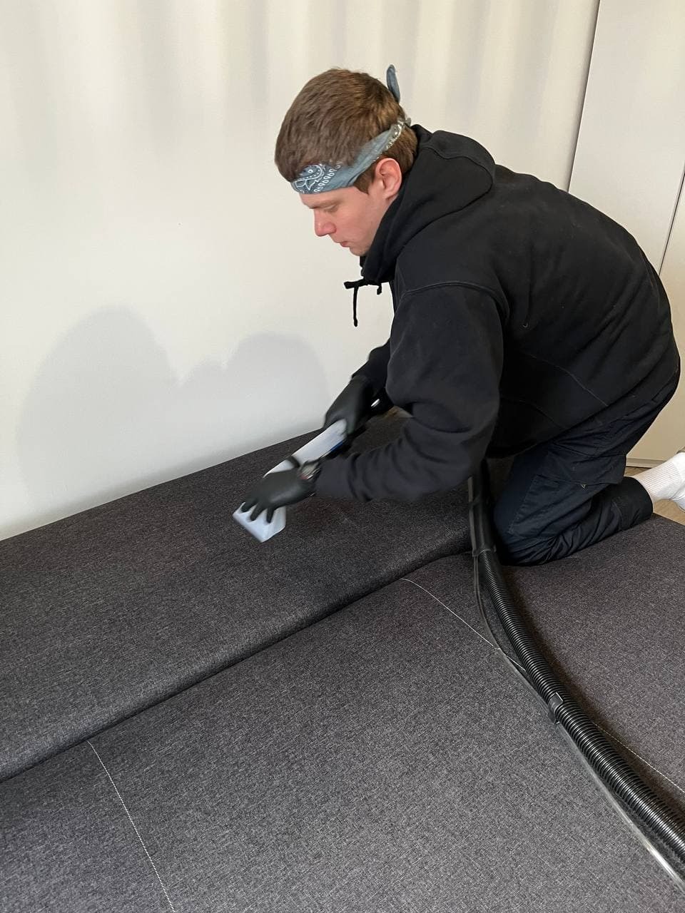 Mężczyzna w czarnej bluzie i rękawicach, z chustą na głowie, klęczy na szarej kanapie i czyści ją za pomocą urządzenia z białą końcówką i wężem ssącym.
