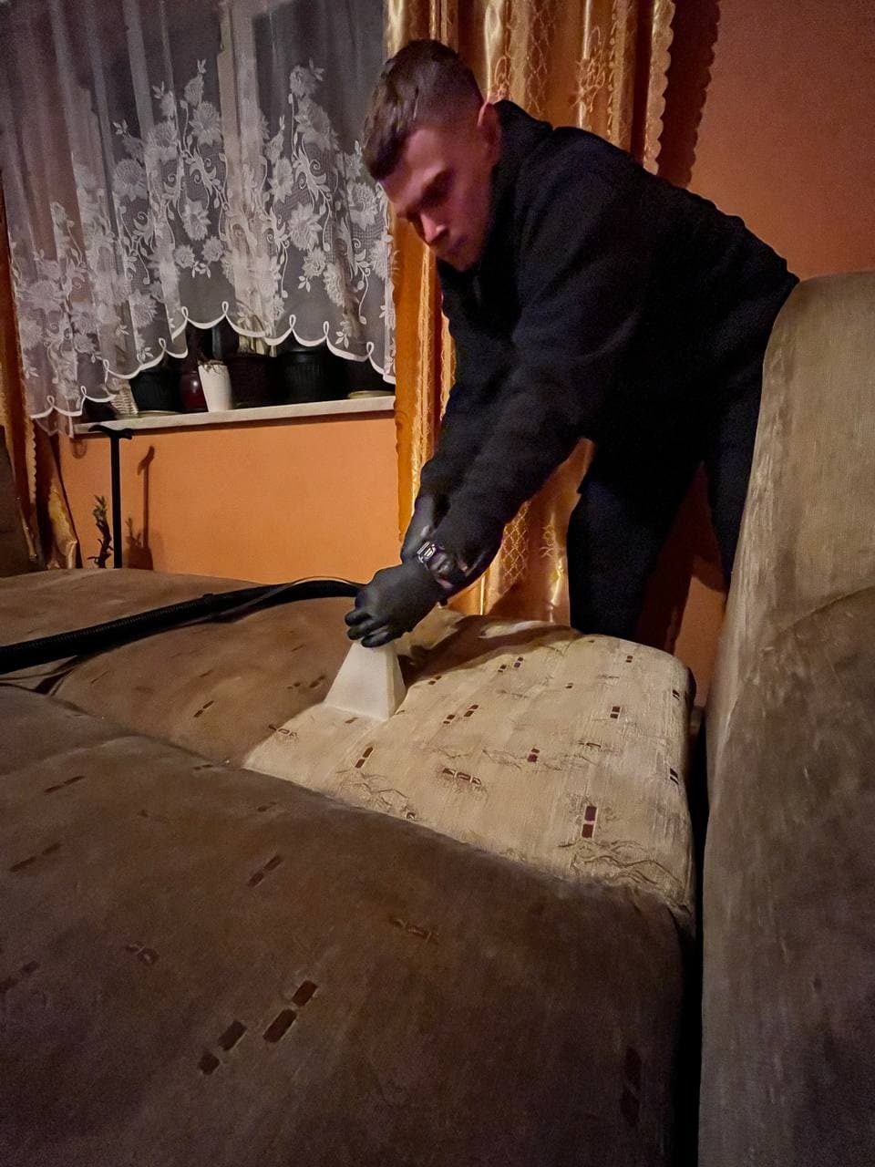 Mężczyzna w czarnej kurtce i rękawicach czyści jasnobrązową kanapę za pomocą odkurzacza piorącego z przezroczystą dyszą, w tle okno z białą firanką i pomarańczowa ściana.
