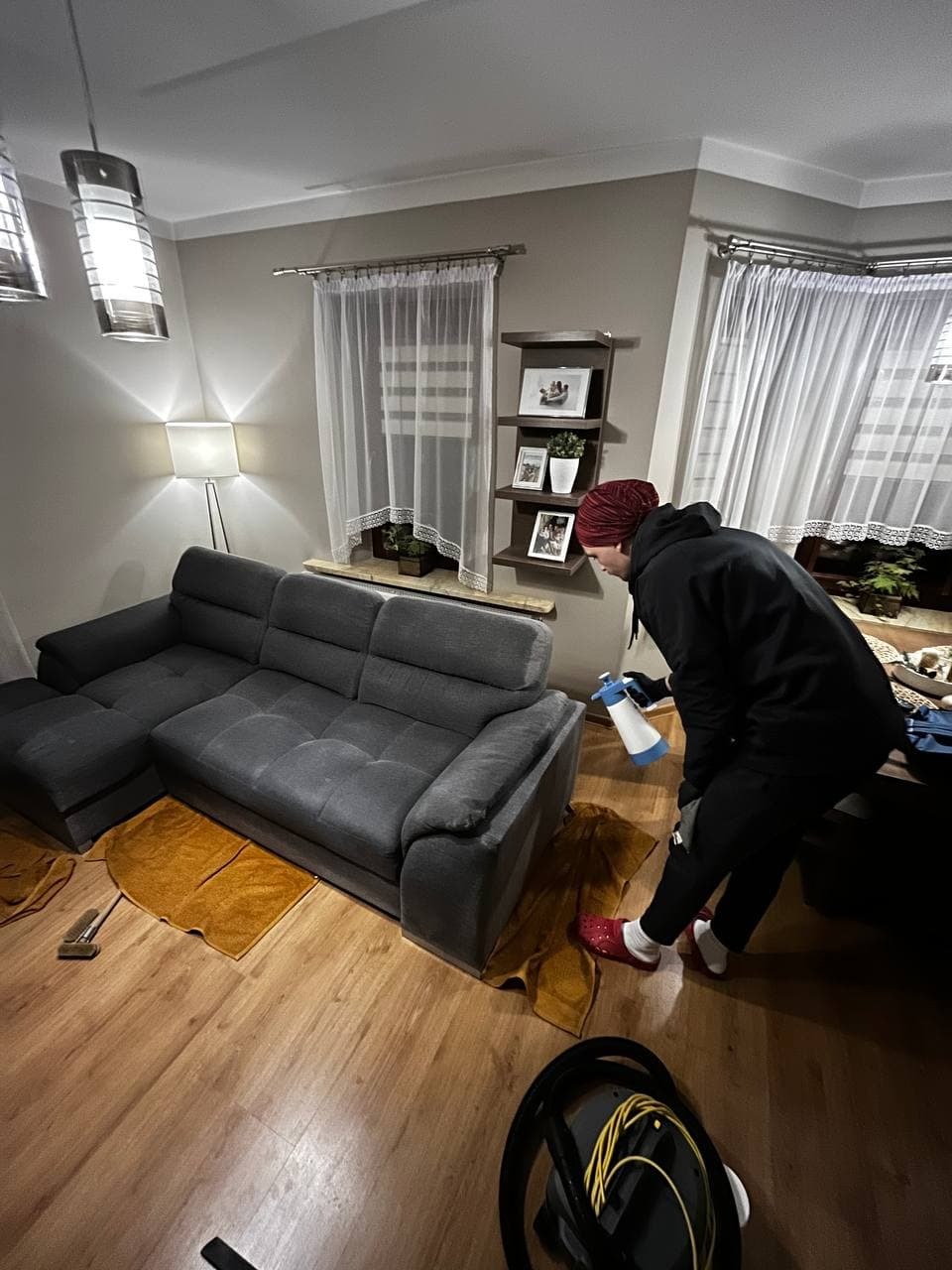 Pracownik w czerwonych klapkach, w trakcie prania narożnika w salonie, używa spryskiwacza z niebieską końcówką, na podłodze leżą ręczniki i wąż od odkurzacza.