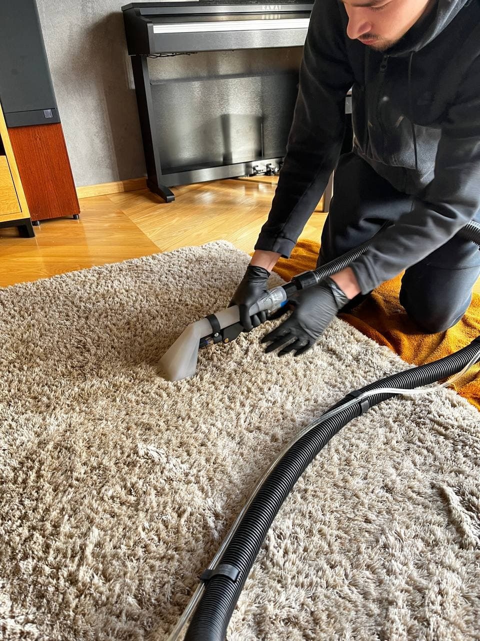 Mężczyzna w czarnych rękawicach czyści beżowy dywan shaggy za pomocą odkurzacza piorącego z przezroczystą końcówką, w tle widoczny czarny fortepian.