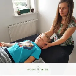 Kobieta delikatnie masuje głowę leżącego dziecka na kozetce w gabinecie w Banino. Dziecko ma zamknięte oczy, a na kozetce leży szary koc.