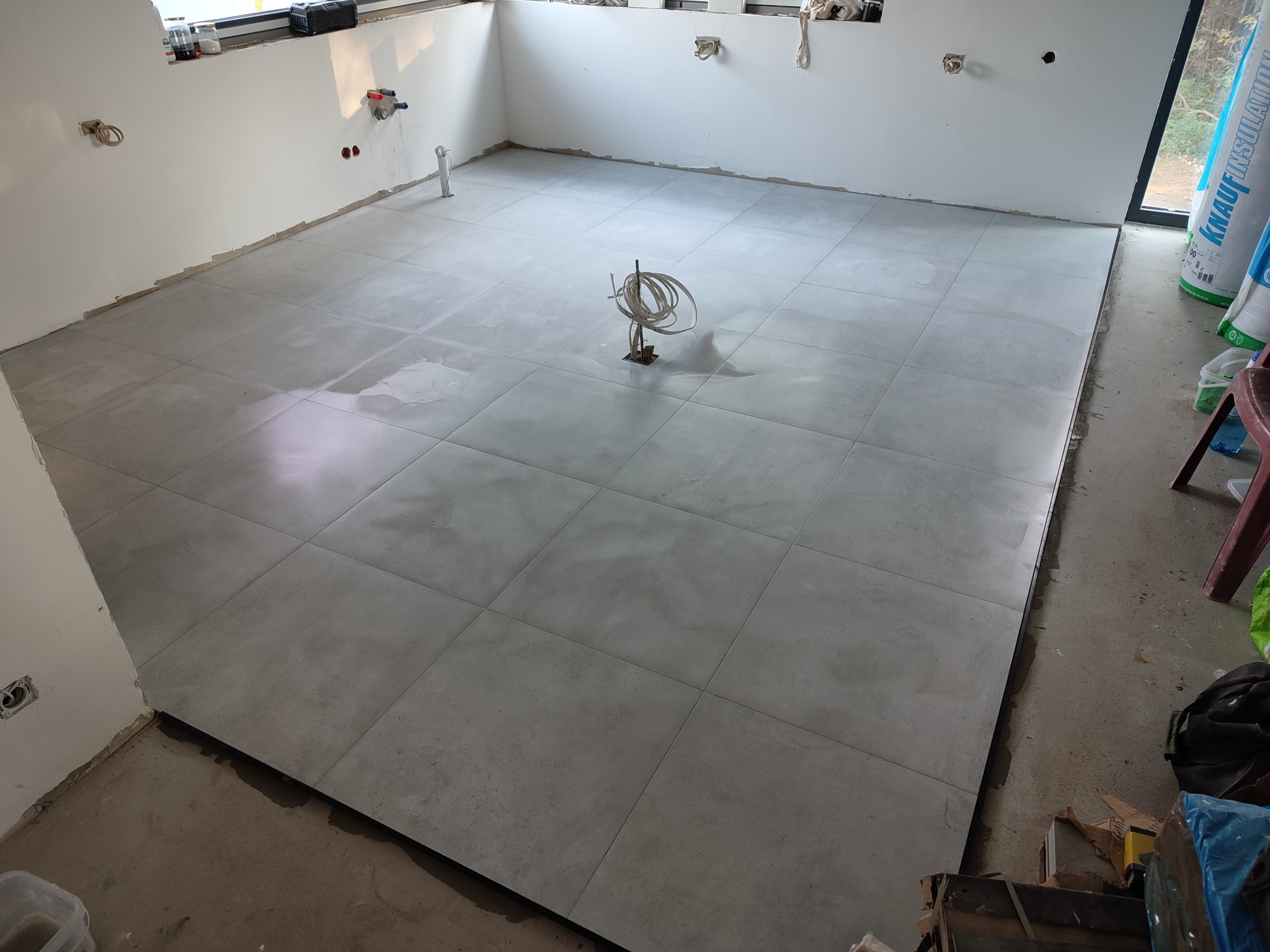 Świeżo ułożone, duże, szare płytki podłogowe w pustym pomieszczeniu, widoczne przewody elektryczne wystające z podłogi i ślady po zaprawie na płytkach, przygotowane do fugowania.