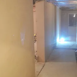 Surowe ściany z nałożoną warstwą tynku w trakcie prac wykończeniowych, widoczne drewniane belki stropowe zabezpieczone folią, fragment podłogi z resztkami materiałów budowlanych.