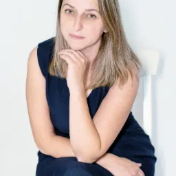 Portret kobiety z blond pasemkami, siedzącej na białym krześle, z ręką podpartą pod brodą, w granatowej sukience na tle białej ściany.