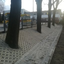 Śląska Manufaktura - Ułożony chodnik z kostki brukowej i ażurowej kostki wokół drzew, palety z kostką w tle, widok z perspektywy ulicy.