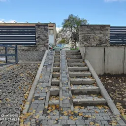 Ułożona z szarej kostki brukowej ścieżka z rampą i schodami, prowadząca do bramy z nowoczesnym, poziomym ogrodzeniem, otoczenie pokryte opadłymi liśćmi.