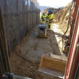 Prace ziemne: Ubijanie gruntu zagęszczarką w wykopie obok izolowanej ściany fundamentowej, widok z kabiny koparki.