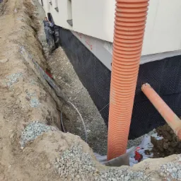 Prace ziemne przy fundamencie budynku: robotnicy montują czarną membranę izolacyjną, widoczny drenaż pionowy z rur karbowanych oraz wykop wypełniony żwirem.