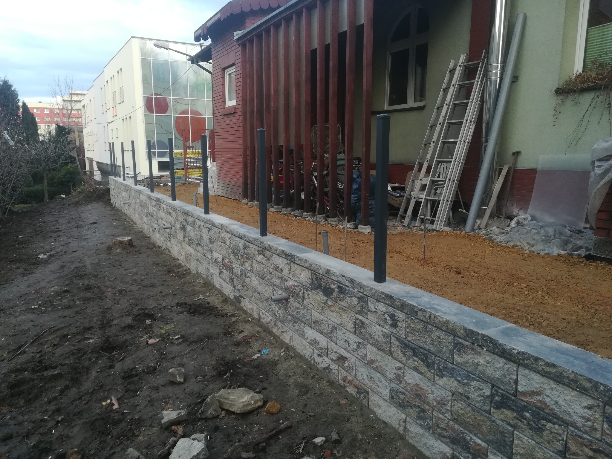 Wzniesiony mur oporowy z szarej, ciosanej kostki na posesji, z czarnymi słupkami i przygotowanym podłożem z piasku; w tle budynek mieszkalny i gospodarczy, drabina oparta o ścianę.