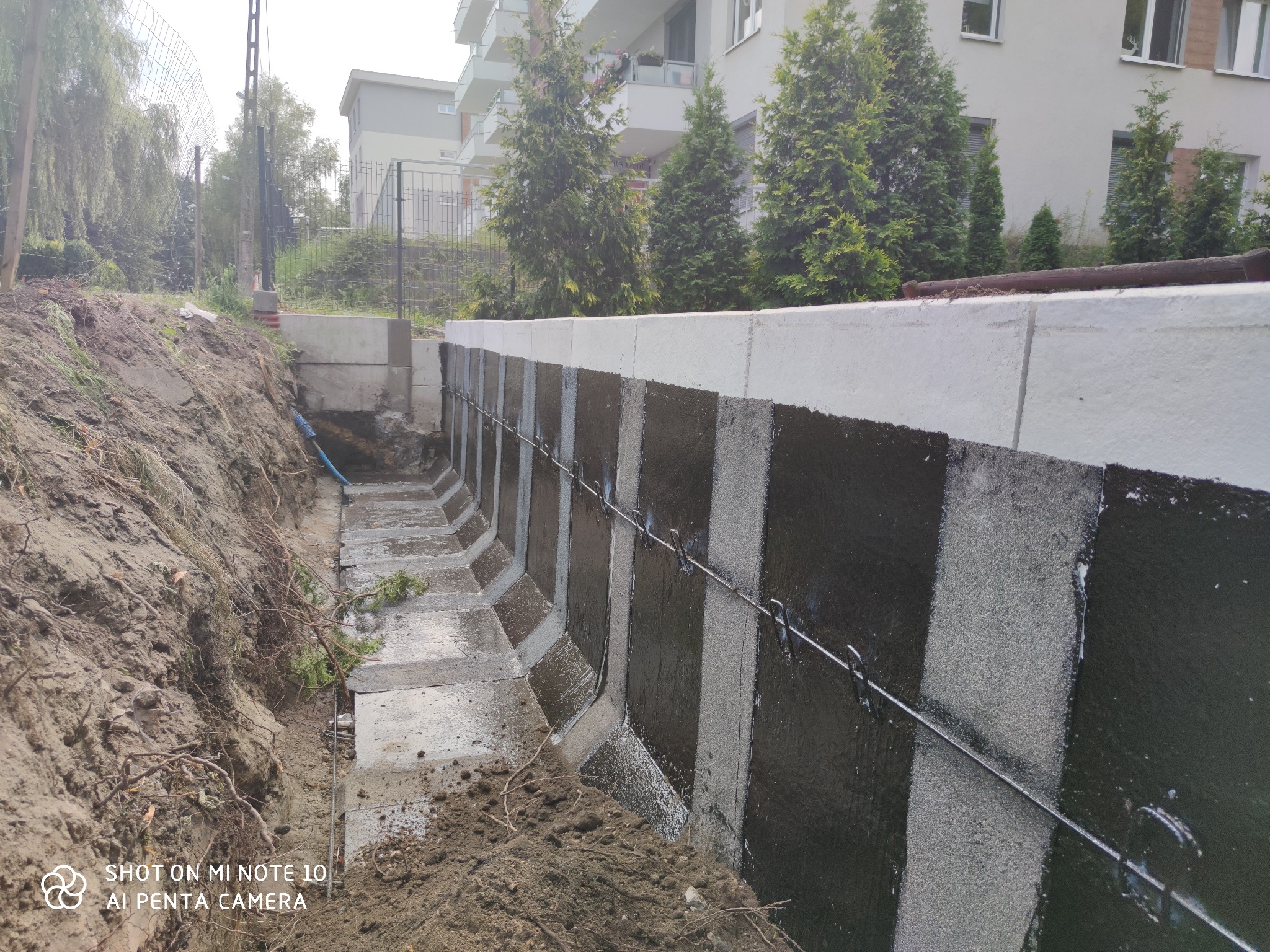 Nowo wybudowany mur oporowy z izolacją pionową, zabezpieczony stalowymi prętami, widoczny wykop z ziemią i fragment budynku w tle.