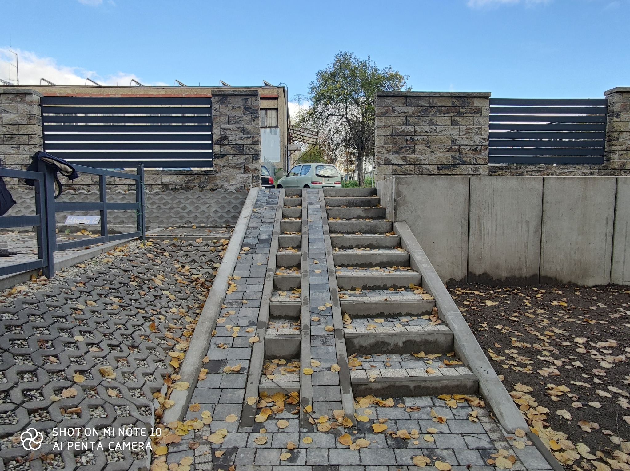 Ułożona z szarej kostki brukowej ścieżka z rampą i schodami, prowadząca do bramy z nowoczesnym, poziomym ogrodzeniem, otoczenie pokryte opadłymi liśćmi.