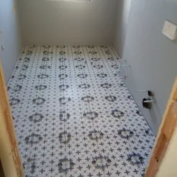 Łazienka w trakcie remontu: ułożone płytki podłogowe z ciemnym wzorem w kształcie kwiatów, krzyżyki dystansowe między płytkami, widoczne rury i otwory w ścianie, przygotowane do dalszych prac...
