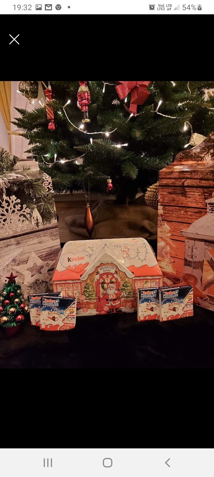 Świąteczny zestaw: puszka w kształcie domku Kinder z kalendarzem adwentowym, czekoladki Kinder i mała choinka, na tle choinki i dekoracji świątecznych.