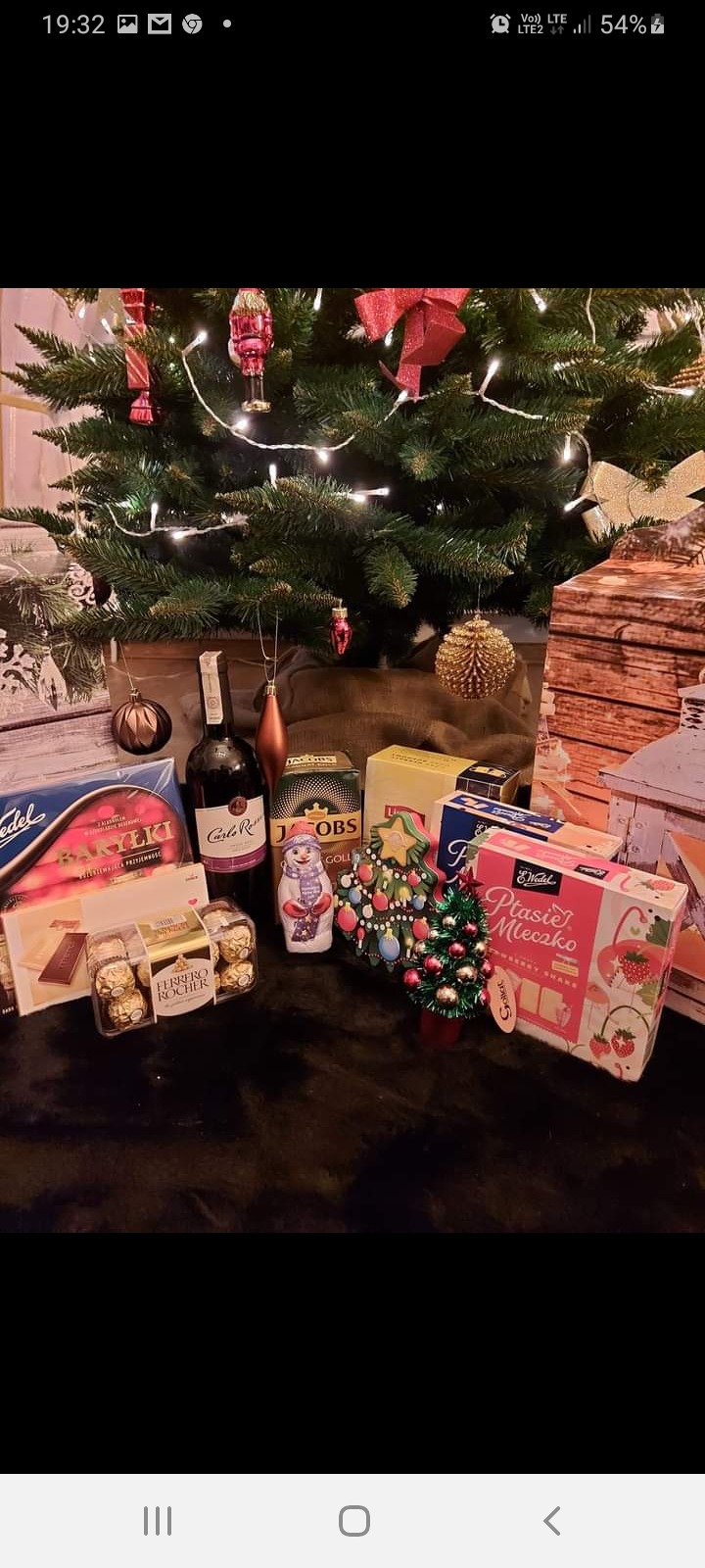 Świąteczny zestaw upominkowy pod choinką, zawierający słodycze, kawę, wino i dekoracje, w tym czekoladowego bałwana i małą choinkę.