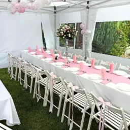 Uroczysty stół w białym namiocie, dekorowany różowym bieżnikiem, kwiatami i balonami, białe krzesła z różowymi wstążkami na trawie.