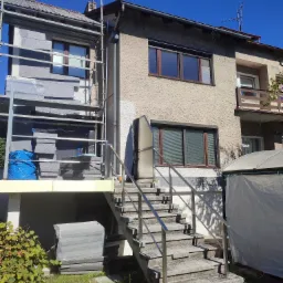 Dom w trakcie ocieplania: styropian grafitowy na rusztowaniu, część elewacji ocieplona, widoczne schody zewnętrzne z metalową barierką i okna z brązowymi ramami.