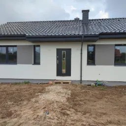 Dom jednorodzinny z jasną elewacją, grafitową stolarką okienną i drzwiami, oraz ciemną dachówką; widoczny fragment terenu przed budynkiem z ziemią i ścieżką z kamieni.