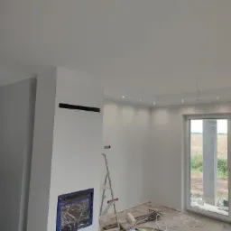 Wnętrze w trakcie wykańczania: świeżo pomalowane ściany na biało, zabezpieczony w folii otwór na kominek, drabina oparta o ścianę, widoczne narzędzia malarskie i kable na podłodze, duże okno...