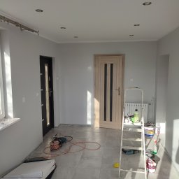 Majster-bud - Wnętrze pokoju po malowaniu ścian na szaro, widoczna drabina z narzędziami malarskimi, zamontowane oświetlenie sufitowe punktowe, drzwi wewnętrzne z przeszkleniami i częściowo rozpakowany sufit...