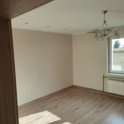 Jasne wnętrze pokoju po remoncie z panelami podłogowymi, widoczne ściany w odcieniach beżu, biały sufit z punktowym oświetleniem, okno z widokiem na zewnątrz i grzejnik pod nim, oraz ozdobny żyrandol.