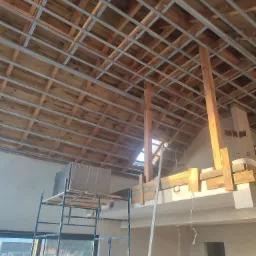 Konstrukcja metalowa pod sufitem podwieszanym, widoczne drewniane elementy więźby dachowej, rusztowanie i fragment okna w tle.