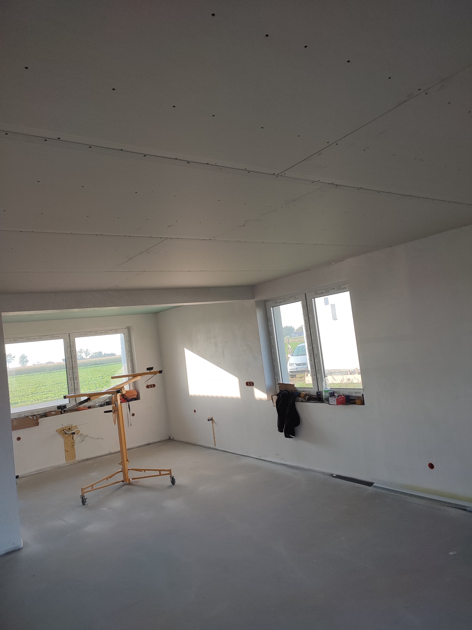 Wnętrze w trakcie wykańczania: sufit pokryty płytami, widoczny podnośnik do płyt, okna z widokiem na zewnątrz, betonowa podłoga.