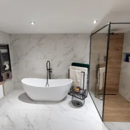Nowoczesna łazienka z marmurowymi płytkami, wolnostojącą wanną z czarną armaturą, kabiną prysznicową typu walk-in z drewnianą ścianą i wnęką z kosmetykami, oraz białymi meblami łazienkowymi...