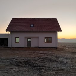 Termo Dom - Nowo wybudowany, otynkowany dom jednorodzinny z czerwoną dachówką, widoczny o wschodzie słońca na niezagospodarowanej działce.