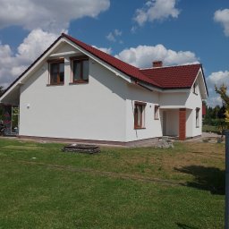 Dariusz -Termo Dom Ostrów Wlkp - Nowoczesny dom jednorodzinny z białą elewacją, brązowymi oknami i czerwoną dachówką, otoczony częściowo zagospodarowanym trawnikiem pod błękitnym niebem.