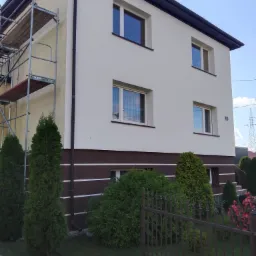 Dwukondygnacyjny dom z częściowo odnowioną elewacją w kolorze kremowym, brązowy pas przy gruncie, widoczne rusztowanie przy jednej ze ścian, ogród z tujami i kwiatami, metalowe ogrodzenie.