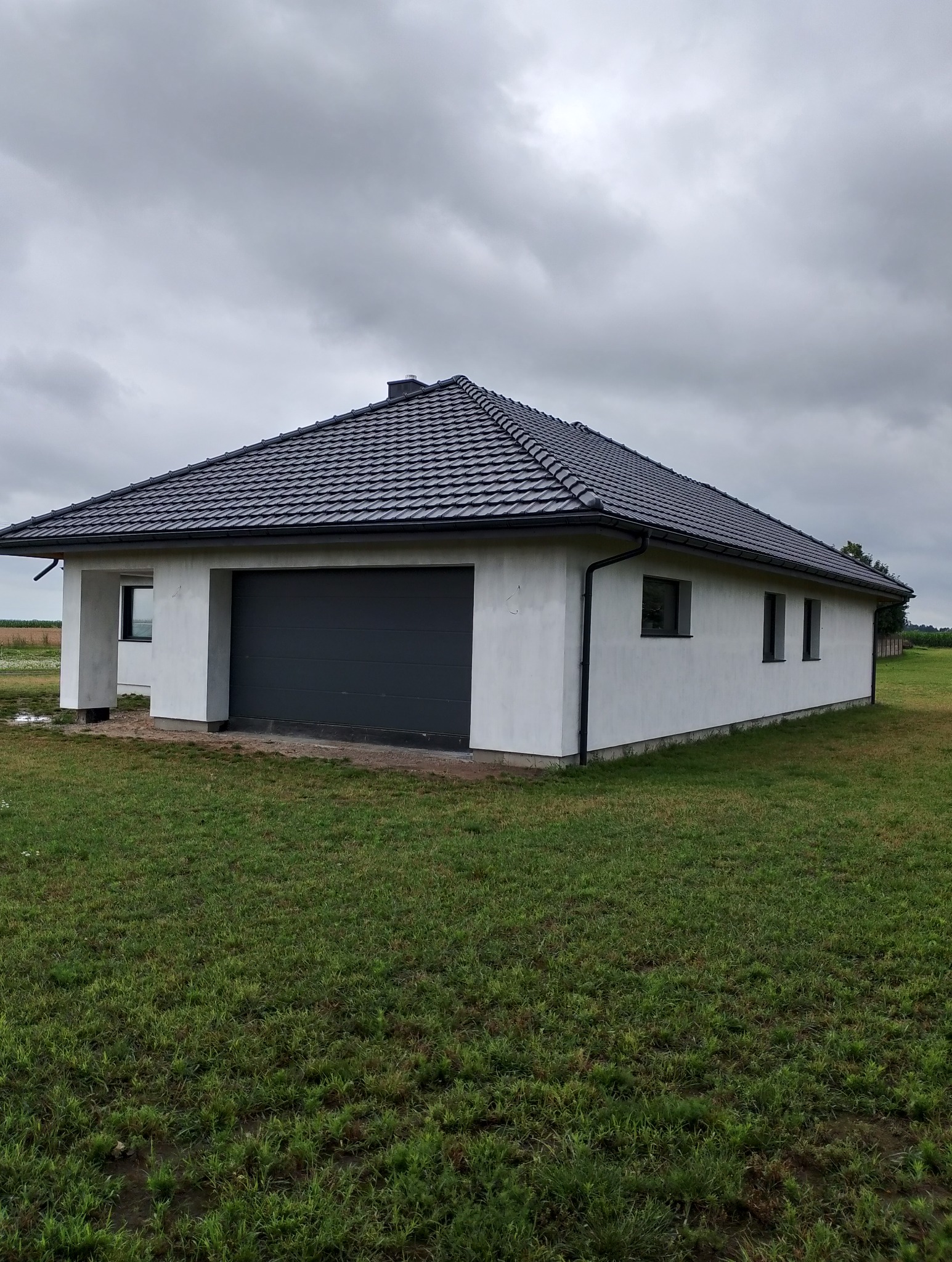 Nowoczesny parterowy dom z garażem, biała elewacja, grafitowa brama garażowa i dachówka, widok z trawnika w pochmurny dzień.