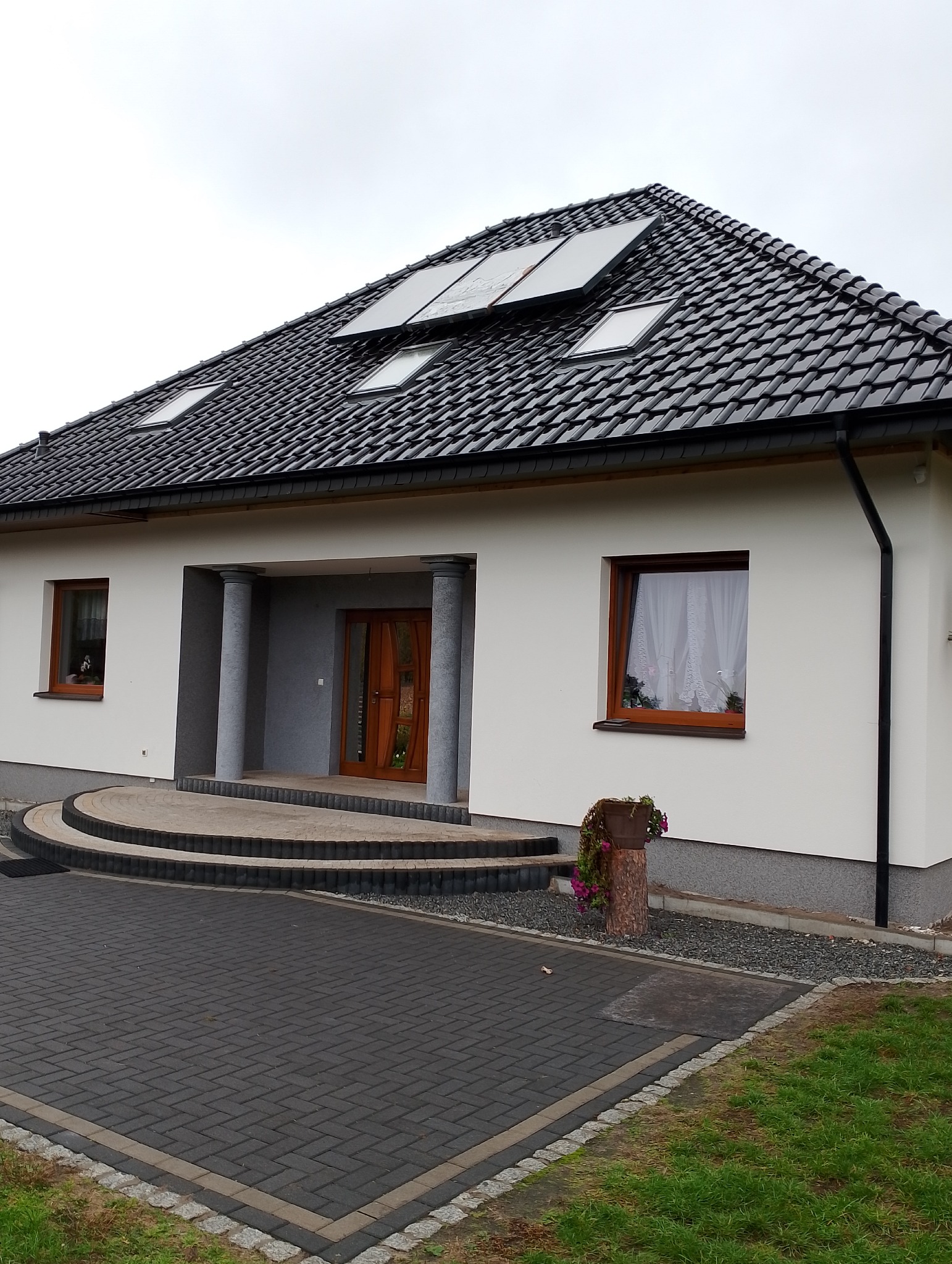 Jednopiętrowy dom z białymi ścianami, ciemną dachówką, panelami słonecznymi i brukowanym podjazdem w pochmurny dzień.