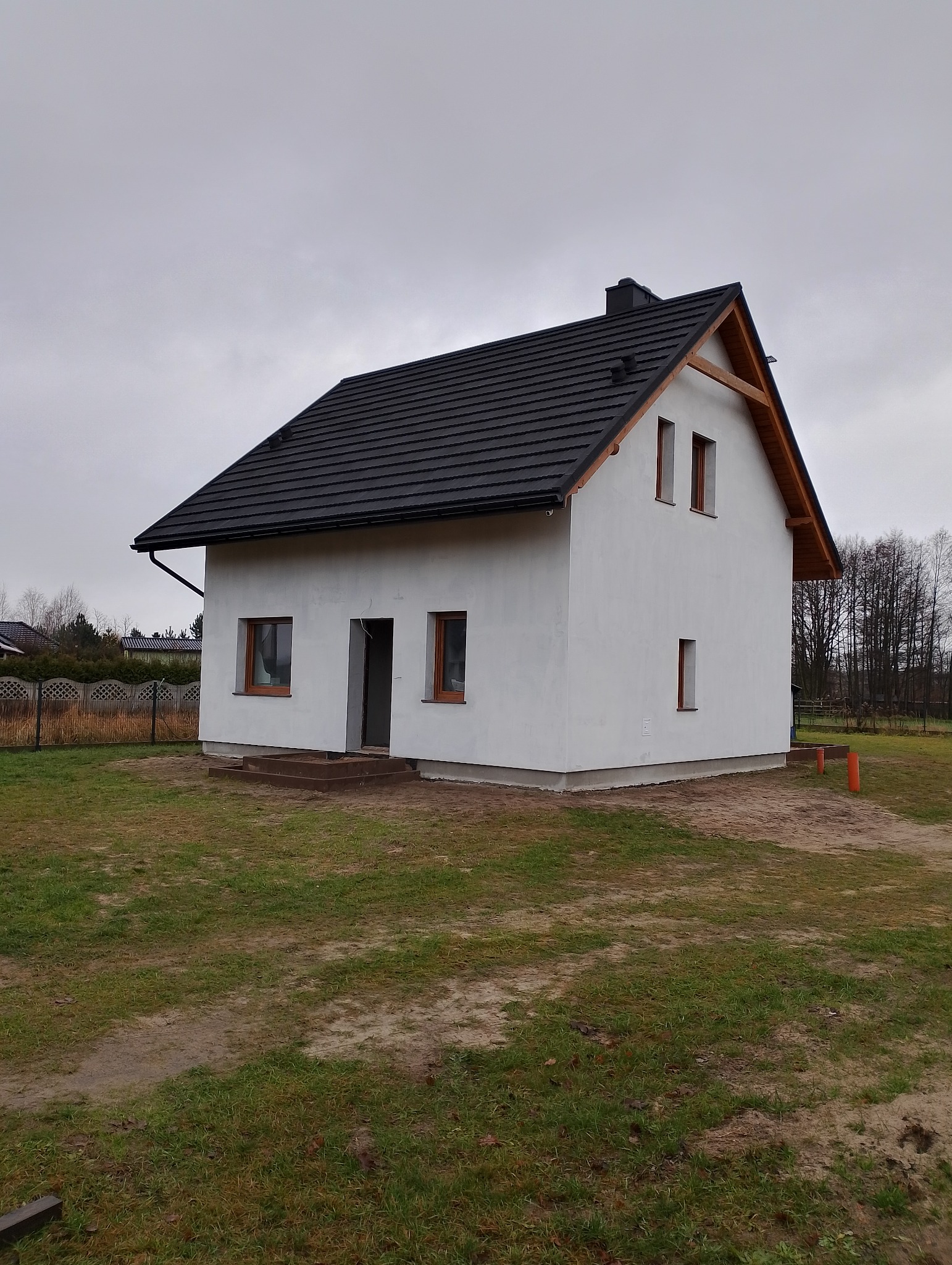 Niewielki, parterowy dom z czarną dachówką i jasną, surową elewacją, widoczne drewniane elementy pod dachem, teren wokół domu częściowo trawiasty, częściowo piaszczysty.