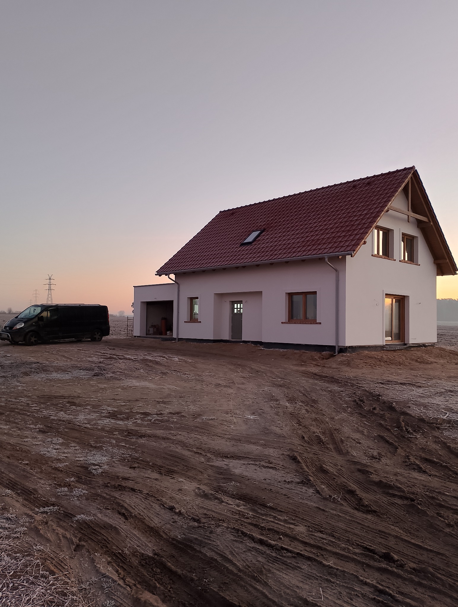 Nowo wybudowany dom jednorodzinny z czerwoną dachówką, białą elewacją i ciemną stolarką okienną, sfotografowany o zmierzchu, z czarnym busem w tle i lekko oszronioną ziemią.