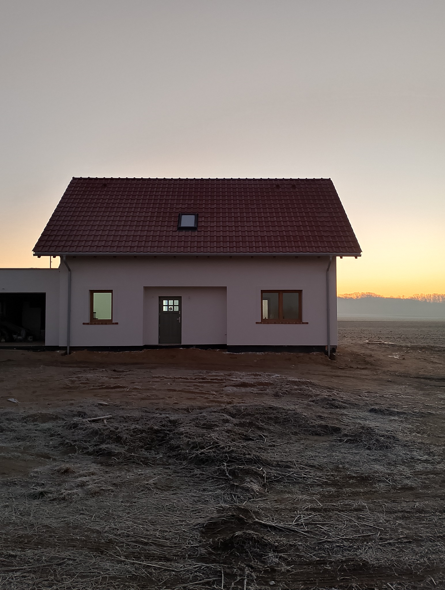 Nowo wybudowany, otynkowany dom jednorodzinny z czerwoną dachówką, widoczny o wschodzie słońca na niezagospodarowanej działce.