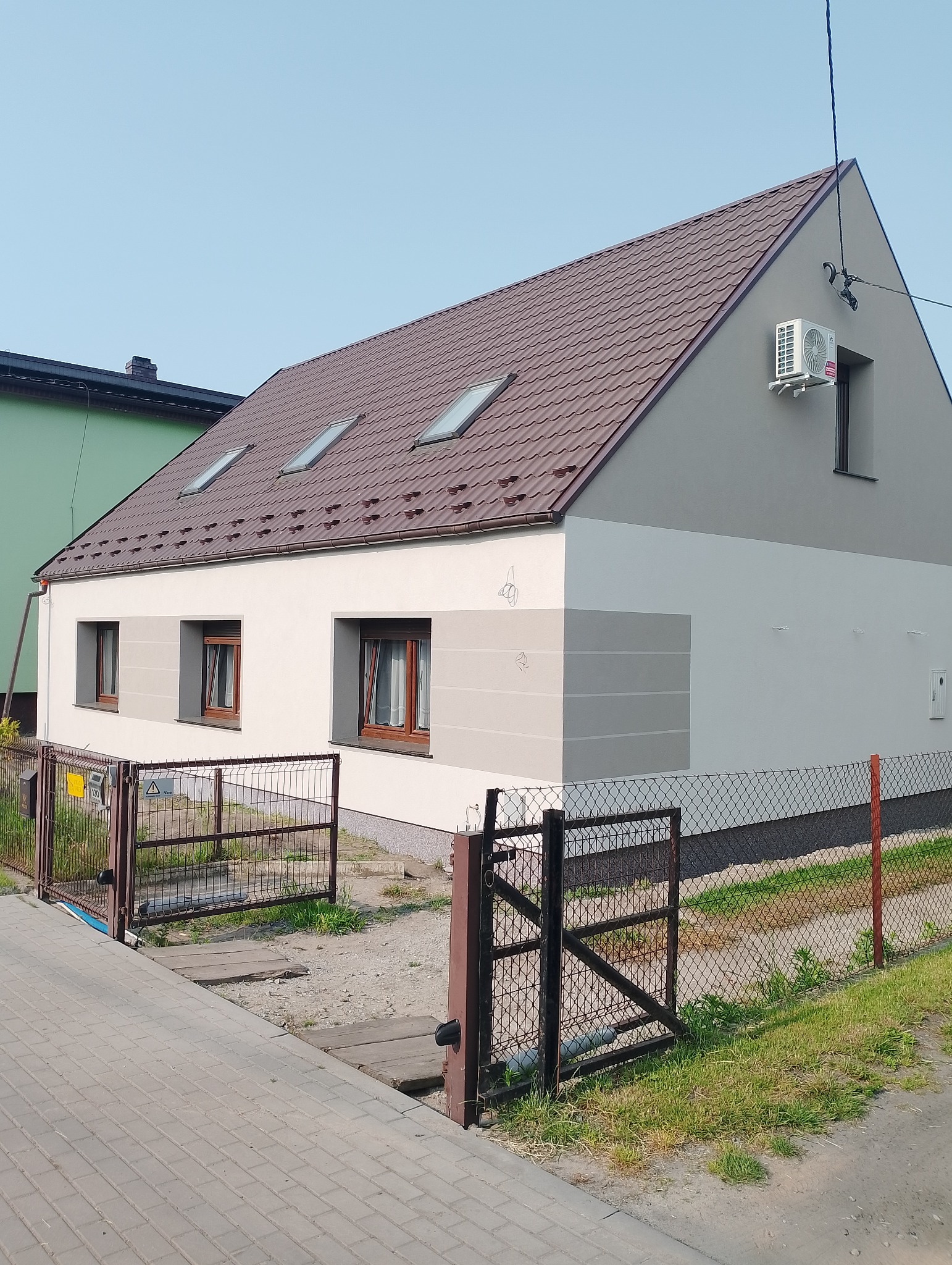 Dwukondygnacyjny dom po renowacji z brązowym dachem i klimatyzacją na szczycie elewacji. Widoczne okna dachowe i elewacja w odcieniach bieli i szarości.