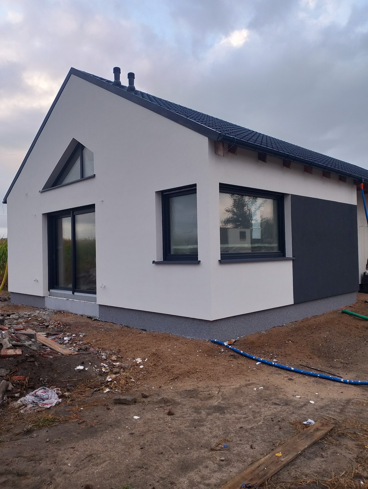 Nowoczesny, parterowy dom z białą elewacją i grafitowym akcentem. Widoczne okna z czarnymi ramami i fragment dachu. Teren wokół domu w trakcie prac wykończeniowych.