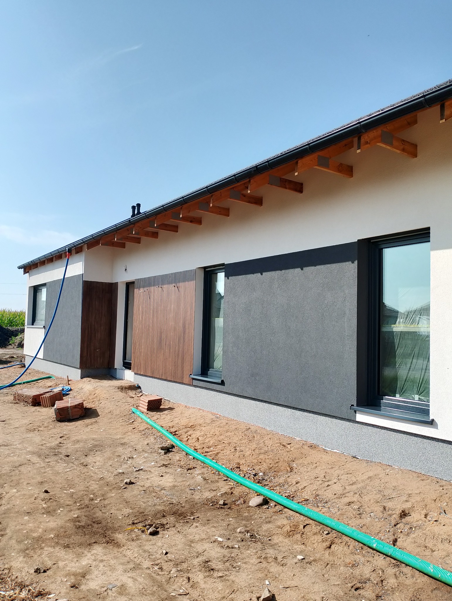 Nowoczesny dom jednorodzinny z elementami drewnianej elewacji i szarym tynkiem. Widoczne okna z folią ochronną, teren wokół domu w trakcie prac wykończeniowych.