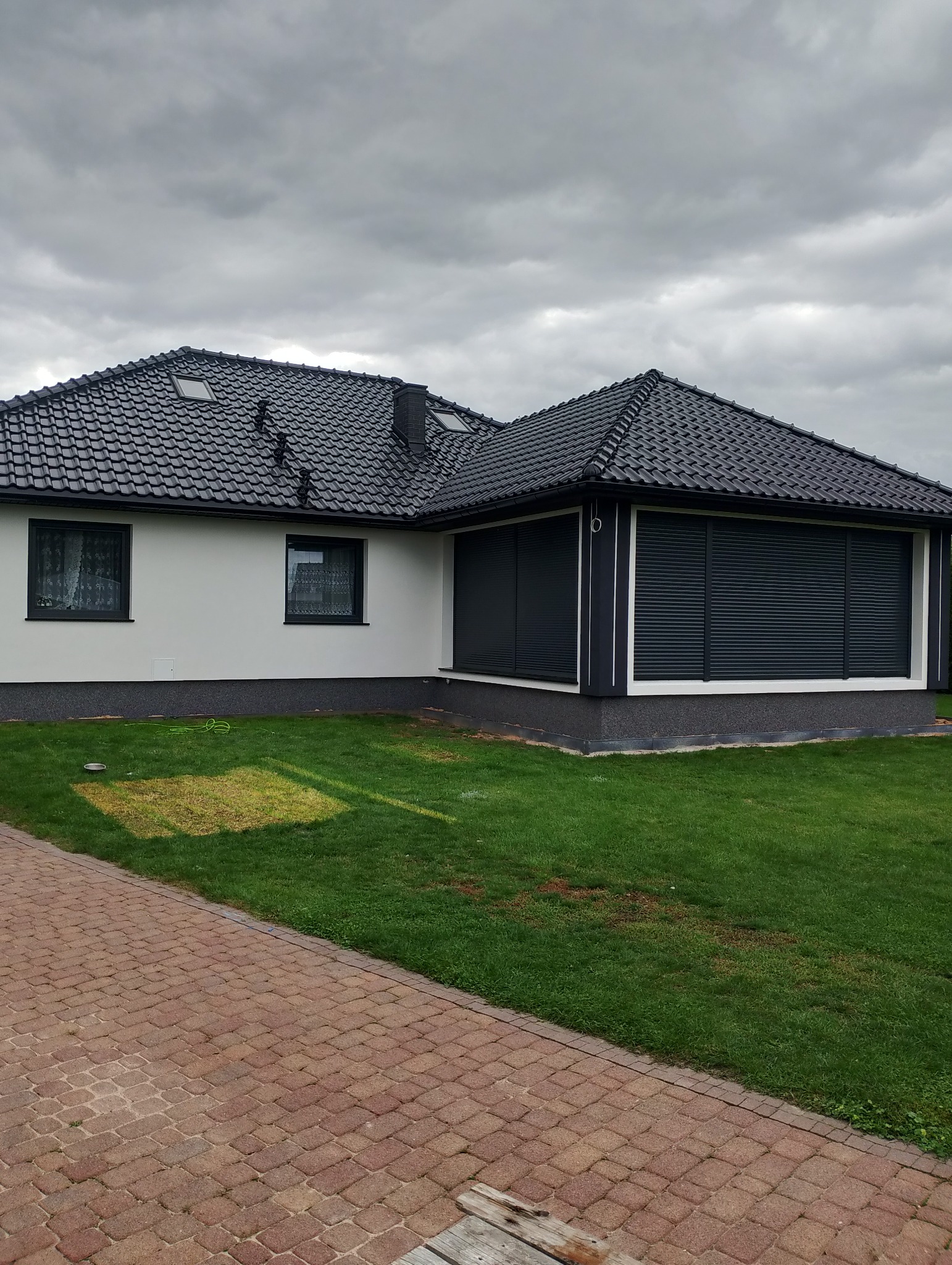 Dom jednorodzinny z ciemną dachówką i jasną elewacją, z zamontowanymi roletami zewnętrznymi w kolorze grafitowym. Widok z ogrodu na część budynku.