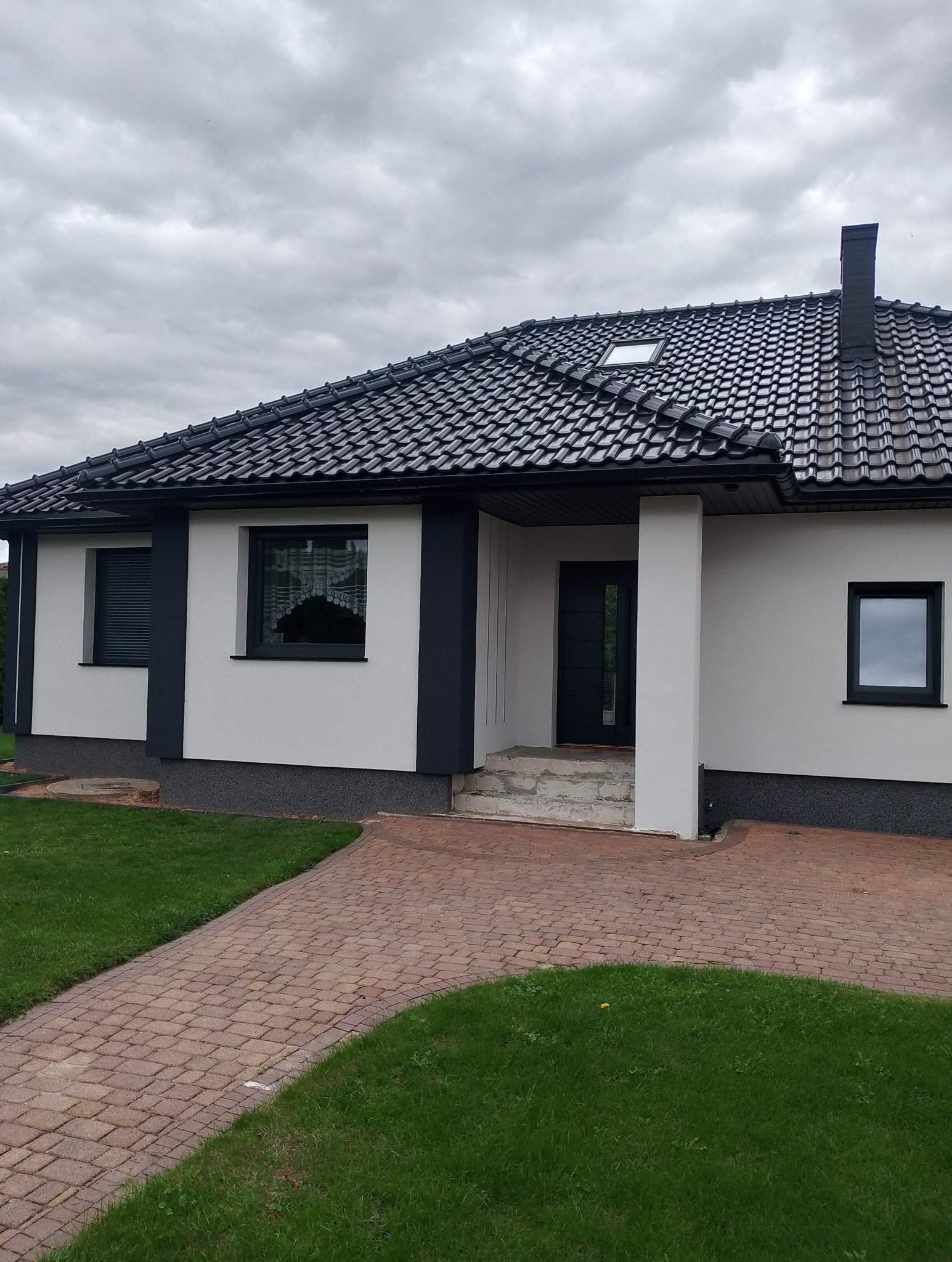 Nowoczesny dom jednorodzinny z białą elewacją i ciemną stolarką okienną oraz dachem z czarnej dachówki ceramicznej. Wejście wyłożone kostką brukową.