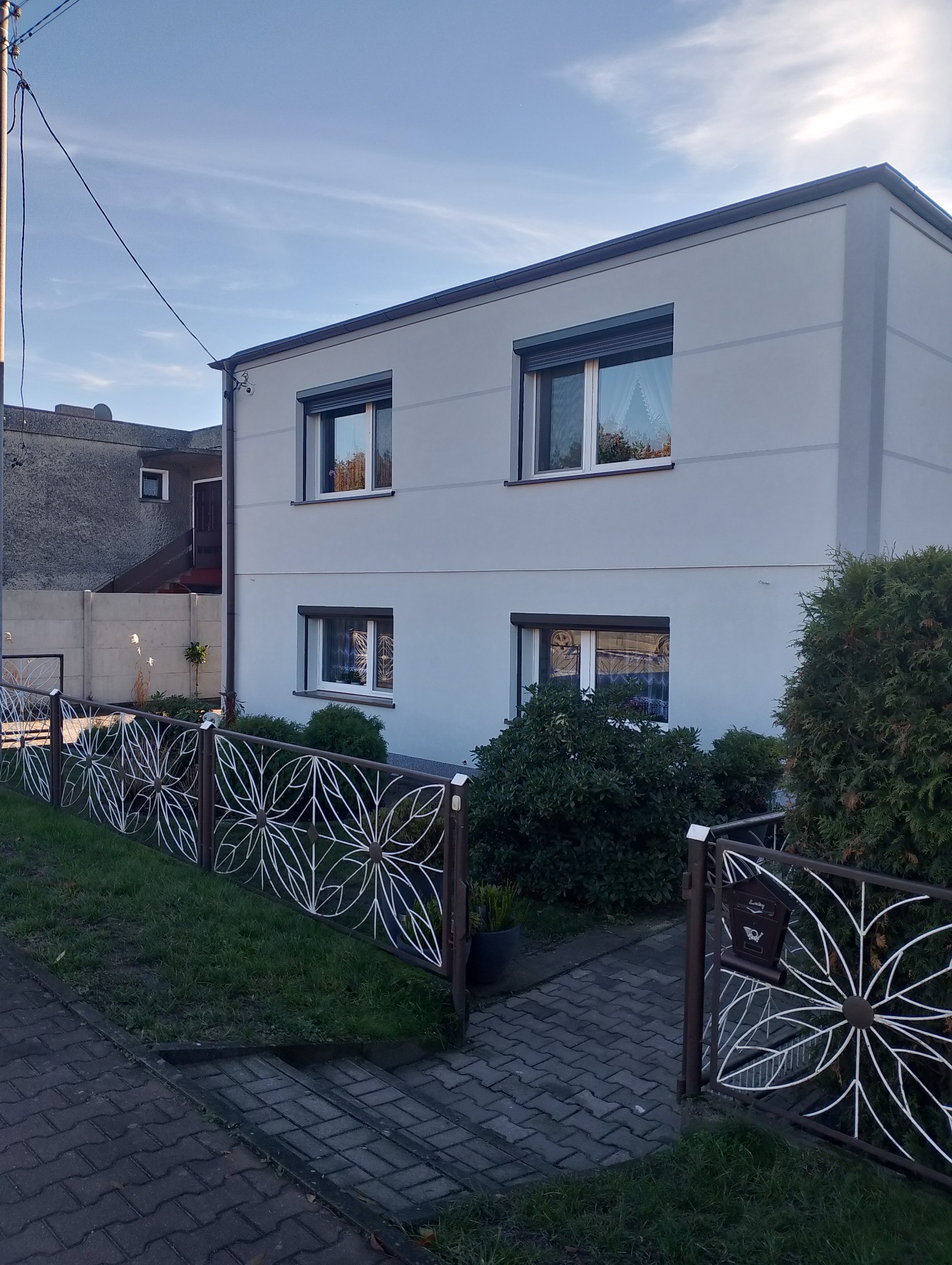 Dwukondygnacyjny dom z jasnoszarą elewacją, widoczny zza ozdobnego, metalowego ogrodzenia. Okna z roletami, ciemny dach. Ujęcie z poziomu chodnika.