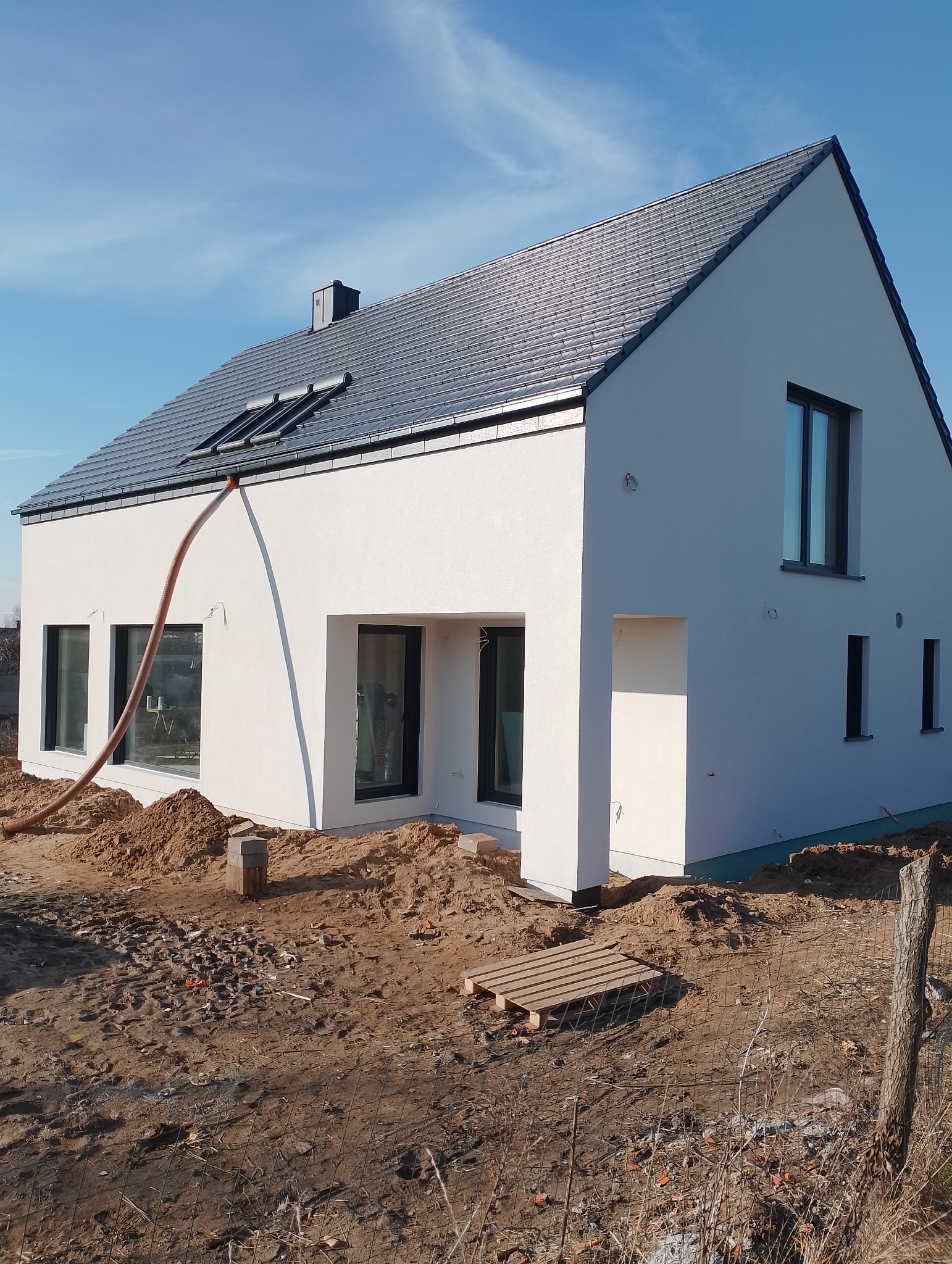 Nowoczesny dom jednorodzinny z białą elewacją i grafitowym dachem, w trakcie prac wykończeniowych. Widoczna rura do wdmuchiwania izolacji.