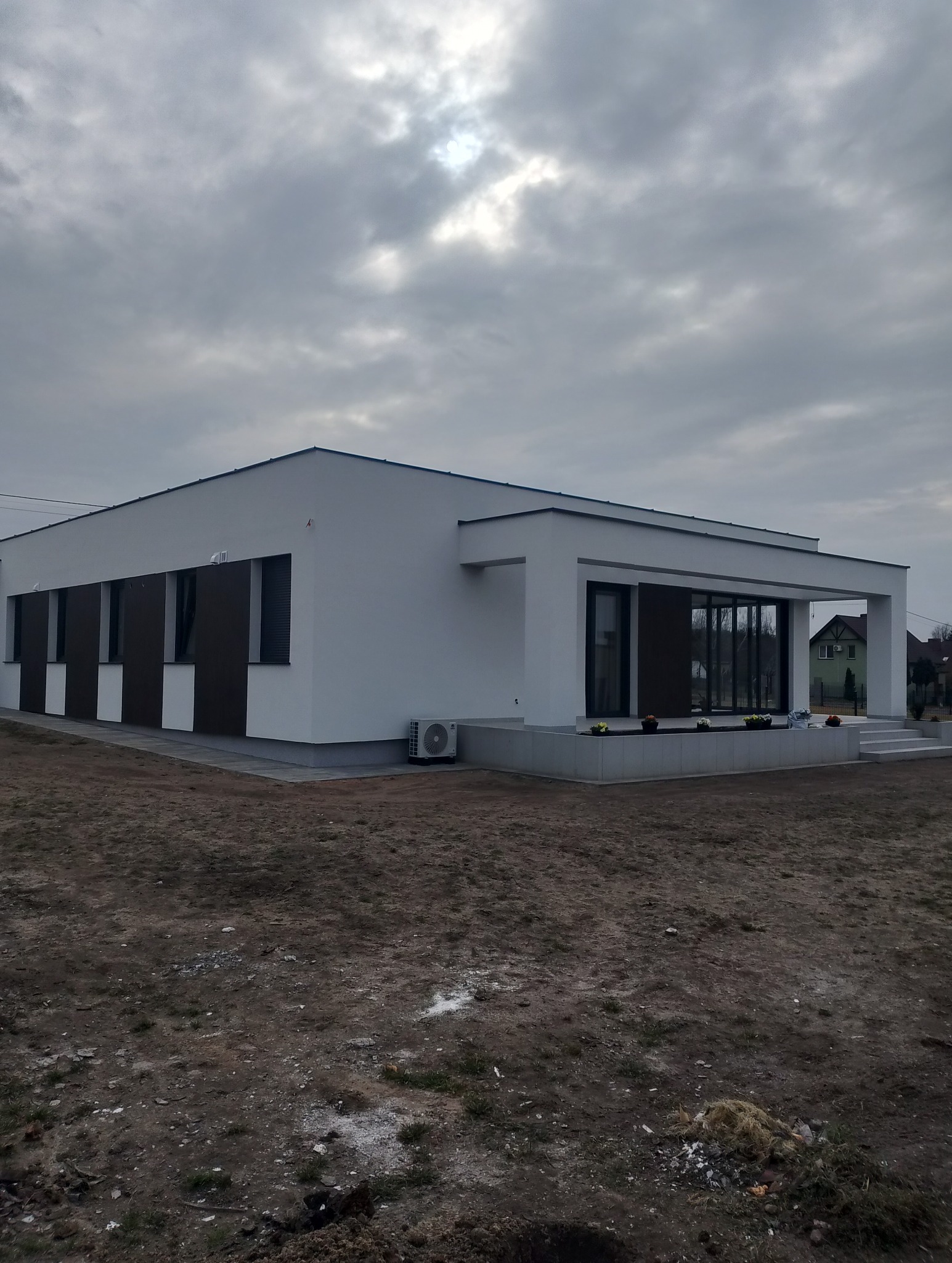 Nowoczesny, parterowy dom z białą elewacją i brązowymi akcentami, widoczny klimatyzator, szara kostka brukowa i pochmurne niebo w tle. Minimalistyczny design.