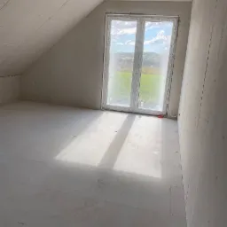 Poddasze w trakcie wykańczania, ściany i skosy obłożone płytami gipsowo-kartonowymi, widoczne łączenia płyt, okno dachowe z widokiem na zielony krajobraz, surowa podłoga przygotowana do dalszych prac.