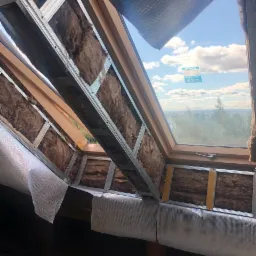 Ocieplenie poddasza wełną mineralną widoczne przez okno dachowe, metalowa konstrukcja szkieletu, folia paroizolacyjna w trakcie montażu, widok na błękitne niebo z chmurami.