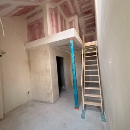 POINT PRIME SOLUTIONS Sp. o.o. - Wnętrze pomieszczenia w trakcie prac wykończeniowych, z widocznymi ścianami i sufitem z płyt gipsowo-kartonowych, częściowo pokrytymi różową masą szpachlową. Wyróżnia się konstrukcja antresoli...